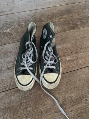 Mörkgröna Converse - Klassiska mörkgröna Converse All Star high tops med vit sula, tå och vita snören. Skorna har rund tå, snörning och den ikoniska loggan på sidan. 