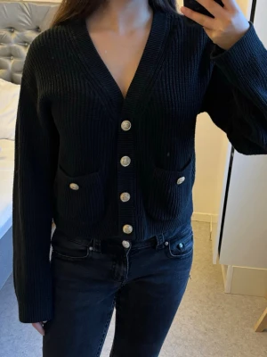 Svart ribbad cardigan med guldknappar - Säljer en svart ribbstickad kofta med croppad passform och snygga guldknappar framtill. Koftan har två fickor på magen, v-ringad hals och långa ärmar. Perfekt att slänga över en topp för en chill men ändå stilsäker look.