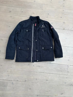 Marinblå Feild jacket från Across the world - Säljer en marinblå feild jacket från Across the world med broderad expeditionstext på bröstet och amerikansk flagga på insidan. Jackan har fyra stora fickor framtill, dragkedja och knappar, samt hög krage. 