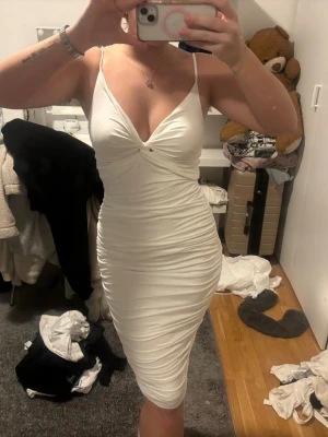 Vit tight kortklänning med rynk - Säljer en vit figurnära kortklänning med smala axelband och snygg rynkdetalj längs sidorna. Klänningen har v-ringning framtill och är tillverkad i ett stretchigt material som sitter snyggt på kroppen. Perfekt för dig som vill ha en stilren och trendig look.