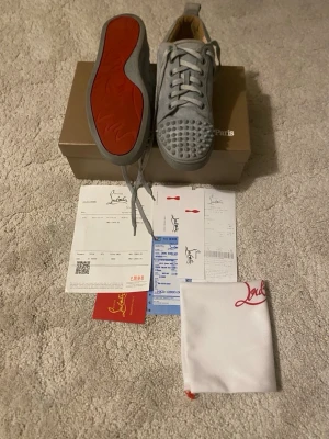 Grå Christian Louboutin sneakers mocka - Säljer ett par grå sneakers från Christian Louboutin i mocka med ikoniska röda sulor och coola nitar på tån. Skorna har snörning och en rund tå. Kommer med originalkartong, dustbag och äkthetsbevis. Perfekta för dig som vill sticka ut med exklusiva detaljer.