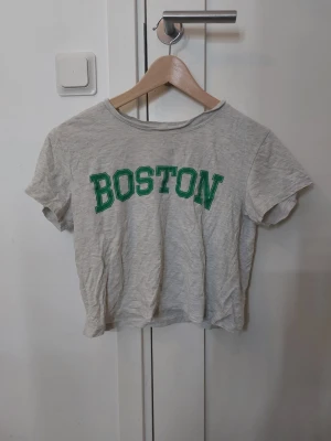 Grå croppad t-shirt Boston Divided - Grå croppad t-shirt från H&M Divided med grön text 'BOSTON' framtill. Klassisk rund halsringning och korta ärmar❣️
