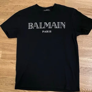 Svart Balmain T-shirt - Säljer en svart t-shirt från Balmain Paris med klassisk vit logga framtill. T-shirten har rund hals och korta ärmar. Tillverkad i mjuk bomull för skön känsla och clean look. Perfekt för dig som gillar stilrena designer och vill ha något från ett lyxigt märke.