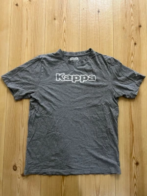 Grå Kappa t-shirt med logga - Snygg grå t-shirt från Kappa i storlek large. Klassisk passform med korta ärmar och rund hals. Stor vit Kappa-logga tryckt på bröstet och diskret logga på ärmen. Perfekt till en avslappnad stil.