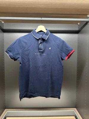 Marinblå pikétröja från Williot - Snygg marinblå pikétröja från Williot med vit piping på kragen och ärmsluten. Ena ärmen har en röd detalj som ger en cool kontrast. Klassisk pikéknäppning med två knappar och diskret logga på bröstet. Perfekt till jeans eller shorts.