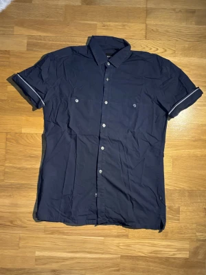 Mörkblå kortärmad skjorta Hugo Boss - Kortärmad mörkblå skjorta från Hugo Boss i slim fit. Skjortan har klassisk krage, vita knappar framtill och två bröstfickor. Tillverkad i mjuk bomull och har diskreta detaljer vid ärmsluten.