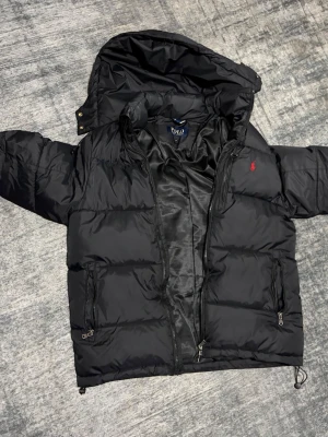 Svart pufferjacka från Polo Ralph Lauren - Säljer en svart pufferjacka från Polo Ralph Lauren med huva och dragkedja. Jackan har två fickor med dragkedja och den klassiska röda Polo-loggan på bröstet. Perfekt för kalla dagar och riktigt snygg till streetwear-stilen.