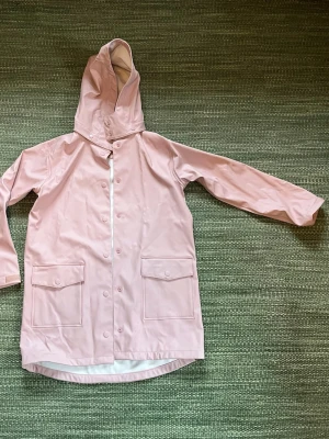 Ljusrosa regnjacka från KappAhl - Snygg ljusrosa ofodrad regnjacka från KappAhl med huva och stora fickor framtill. Jackan har tryckknappar längs hela framsidan och är tillverkad i ett vattenavvisande syntetmaterial. Perfekt för regniga dagar och enkel att matcha med din outfit.