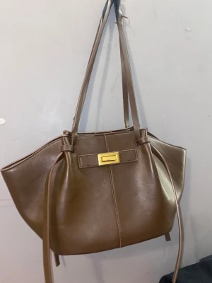 Brun axelväska i skinn med gulddetalj - Möts upp bara då de enklast för mig. Snygg brun axelväska i skinn med långa remmar och en stilren guldfärgad spänndetalj framtill. Väskan har en mjuk, rundad form och dekorativa knytband på sidorna. Perfekt för dig som vill ha en klassisk men trendig look.