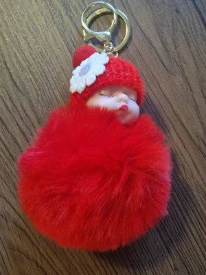 Nyckelring med sovande baby och röd fluffboll - Söt nyckelring med en sovande baby i en röd stickad mössa med en vit blomma. Babyn är omgiven av en stor, röd fluffboll. 11 cm hög. Bredd 8 cm. Perfekt accessoar för väskan eller nyckelknippan.
