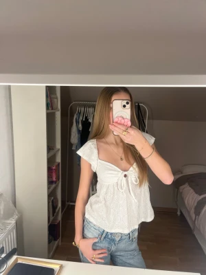 Vit blus med knyte fram  - Superfin vit blus med broderade blommor och knyt framtill. Toppen har v-ringning, korta ärmar och en luftig passform som är perfekt för varma dagar. Söt och romantisk stil som passar till jeans eller kjol.