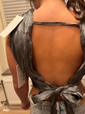 Silvrig topp - Säljer en  silvrig topp från NA-KD med breda axelband, djup v-ringning och öppen rygg. Alldrig använd med prislapp kvar!!