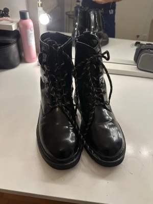 Svarta boots med snörning och dragkedja - Säljer ett par glansiga svarta boots från Pepper med snörning och dragkedja på sidan. Skorna har rund tå, platt sula och är fodrade med mjukt fuskpälsfoder för extra värme. Perfekta för kyligare dagar och ger en cool look till din outfit.