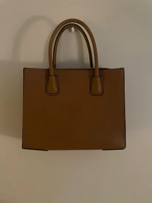 Brun handväska i skinnimitation - Snygg fyrkantig handväska i brun skinnimitation med två rejäla handtag och dragkedja upptill. Väskan har flera fack inuti, inklusive ett med dragkedja, vilket gör den perfekt för att hålla ordning på dina grejer. Enkel och stilren design som passar till det mesta.