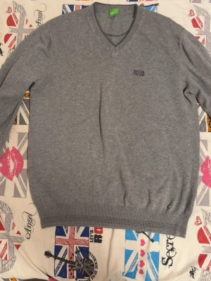 Grå V-ringad tröja från BOSS - Säljer en stilren grå V-ringad tröja från BOSS med diskret logga broderad på bröstet. Tröjan har långa ärmar och ribbade muddar vid ärmslut och nederkant. Perfekt till lager på lager och funkar året runt.