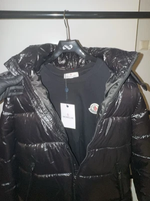 Svart jacka och T-shirt från Moncler - Säljer en svart pufferjacka från Moncler med glansig finish och klassisk logga på ärmen. Och en svart T-shirt från Moncler med klassisk logga på bröstet i topp kvalité. Jackan har dragkedja framtill, fickor med både dragkedja och knapp, samt en stor huva. Perfekt för kalla dagar och riktigt snygg till streetwear-stilen.