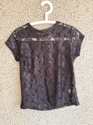 Svart spetsblus med blommönster - Säljer en svart blus i genombruten spets med blommigt mönster. Toppen har rund halsringning och korta ärmar. Perfekt att styla över ett linne eller bh för en trendig look.