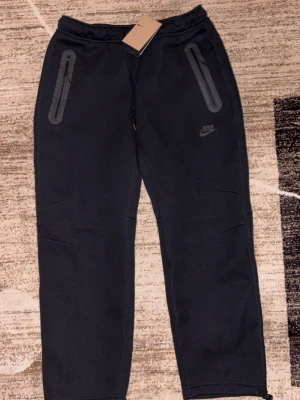 Svarta mjukisbyxor från Nike, storlek L - Etikett finns.Svarta mjukisbyxor från Nike i storlek L. Byxorna har dragkedjor på fickorna och justerbara benslut. Tillverkade i mjukt material som passar perfekt för chill eller träning. Nike-logga på ena benet för clean look.