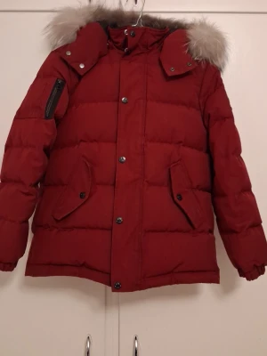Röd Moose Knuckles  - Säljer en röd pufferjacka från Moose Knuckles med stor huva och fuskpäls. Jackan har flera fickor, dragkedja och knappar framtill samt en snygg logga på ärmen. Perfekt för kalla vinterdagar och riktigt varm. Helt ny ca 5000-6000 säljer nu för 1500. St på jackan är M skulle samt säa att jackan passar en åldee från 10-12. Pris kan absolut diskutera