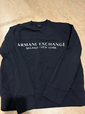 Mörkblå sweatshirt från Armani Exchange - Säljer en mörkblå sweatshirt från Armani Exchange med vit logga och texten 'Milano/New York' framtill. Tröjan har rund halsringning och långa ärmar. Perfekt för dig som gillar stilrena och klassiska plagg med en modern touch.