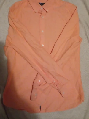 Persikofärgad skjorta från Easy Wear - Persikofärgad skjorta från EAST WEST med klassisk krage och vita knappar framtill. Skjortan har lång ärm och är tillverkad i ett mjukt bomullstyg. Perfekt för dig som vill ha en enkel men snygg skjorta med clean look.