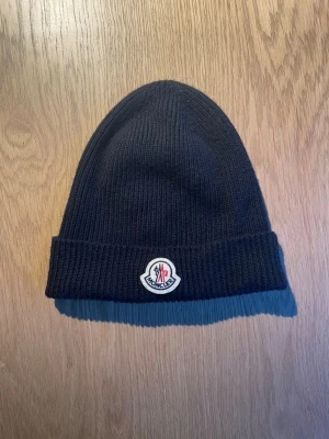 Marinblå Moncler mössa i ull - Nypris är 2500kr. Har kvitto. Säljer en marinblå ribbstickad mössa från Moncler med klassisk uppvikt kant och Moncler-logga framtill. Mössan är gjord i mjuk ull och har en enkel, stilren design som passar perfekt till kalla dagar.