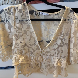 Blommig spetsblus från Zara - Säljer en transparent, beige blus från Zara med blommigt spetsmönster, knappar och volanger. Så sjukt fin!