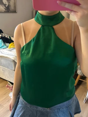 Grön halterneck linnetopp i satin - Säljer en grön halterneck linnetopp i glansigt satinliknande material. Toppen har hög krage och är ärmlös med en snygg, slät finish. Perfekt att styla med jeans eller kjol för en trendig look.