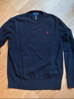 Stickad Ralph Lauren tröja  - Säljer en mörkblå långärmad tröja från Polo Ralph Lauren med klassisk röd logobrodering på bröstet. Tröjan har rund hals och ribbade muddar vid ärmslut och nederkant. Perfekt för dig som gillar stilrena och tidlösa plagg.
