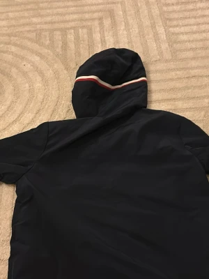 Mörkblå vindjacka från Moncler - Säljer en mörkblå vindjacka från Moncler med huva och snygga rödvita ränder. Jackan har dragkedja framtill, snörning i huvan och två fickor med tryckknappar. Klassisk Moncler-logga på ärmen. Perfekt för den kommande vår/sommar. (Original pris 3499) 