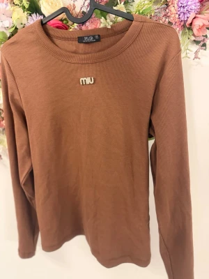 Brun ribbad långärmad tröja NLG - Säljer en brun ribbad långärmad tröja från NLG Collection med broderad text 'miu' framtill. Tröjan har rund halsringning och är i ett mjukt, stretchigt material som sitter skönt på kroppen.