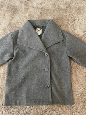 Grå kappa från Gina Tricot - Stilren grå kappa från Gina Tricot med bred krage och tre knappar framtill. Klassisk rak passform och mjukt material som känns skönt mot huden. Perfekt för dig som vill ha en enkel men snygg jacka till höst och vinter.tar gärna emot bud och skriv om ni har frågor💕😊