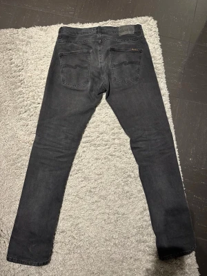 Nudie Jeans - Säljer ett par svarta jeans från Nudie Jeans i storlek 31/32 grim Tim. De har klassisk femficksdesign, raka ben och diskret logga på bakfickan. Jeansen är i ett mjukt denimtyg och har en snygg tvättad finish som ger en avslappnad vibe.