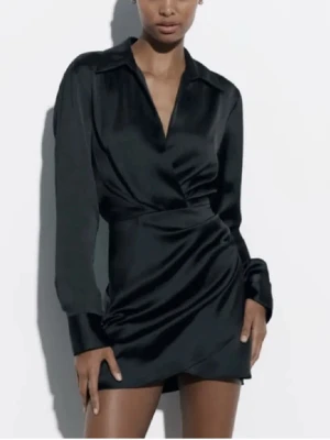 Svart Zara klänning i satin - Snygg svart kort omlottklänning i satin med lång ärm och djup v-ringning. Klänningen har en lätt glansig finish och markerad midja som ger en elegant siluett. Perfekt för dig som vill sticka ut med stil. Klänningen har aldrig används. 