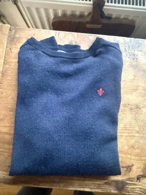 Mörkblå merinoulltröja Morris M - Klassisk mörkblå tröja från Morris i 100% merinoull. Tröjan har rund hals, ribbade muddar och en liten röd broderad logga på bröstet. Perfekt för dig som gillar stilrena och tidlösa plagg med en snygg detalj.