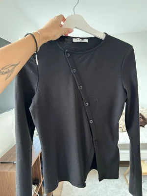 Svart asymmetrisk topp från NA-KD - Säljer en svart långärmad topp från NA-KD med unik asymmetrisk knäppning framtill. Perfekt för dig som gillar stilrena plagg med en twist. Använd fåtal gånger