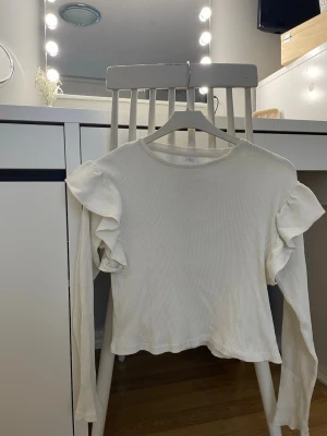 Vit ribbad topp med volang från Zara - Säljer en vit långärmad topp från Zara i ribbad bomull. Toppen har volangdetaljer på axlarna som ger en extra touch och en enkel rund halsringning. Perfekt att styla med jeans eller kjol för en clean look.