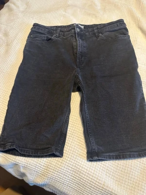 Svarta jeansshorts med fem fickor - Säljer ett par svarta jeansshorts med klassisk femficksdesign och bälteshällor. Shortsen har dragkedja och knapp framtill samt raka ben som slutar strax ovanför knät. Perfekta för en avslappnad stil i sommar.