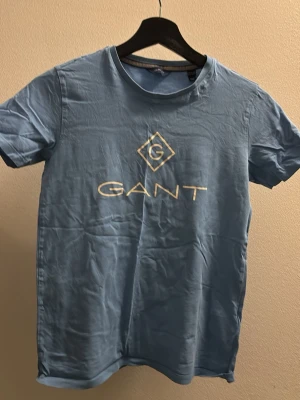 Blå GANT t-shirt med logga - Ljusblå t-shirt från GANT med stor vit logga tryckt på bröstet. Klassisk rund halsringning och korta ärmar. Tillverkad i mjuk bomull.