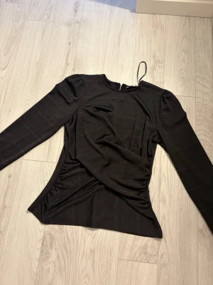 Svart långärmad topp från C - Svart långärmad topp från Cubus med rund halsringning och dragkedja i nacken. Toppen har lätt puffade axlar och draperad effekt i sidan som ger en snygg siluett. Tillverkad i ett mjukt, stretchigt material som känns bekvämt mot huden. Använt två gånger.