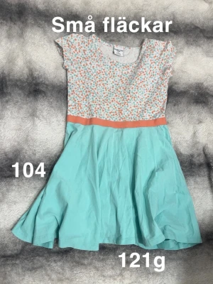 Blommig klänning  - Söt klänning från Polarn o pyret i storlek 104. Överdelen har ett blommigt mönster i orange och turkos på vit botten, med en kontrasterande orange linje i midjan. Kjoldelen är enfärgat ljusblå. 🌸 Små fläckar. Storlek 104