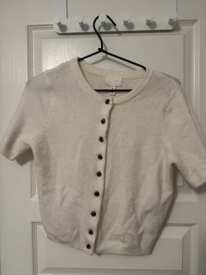 Kortärmad kofta  - Kortärmad vit kofta från H&M i mjukt stickat material. Rund halsringning och svarta/mörkbruna knappar framtill. Perfekt att slänga över ett linne!😽💋