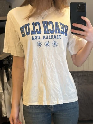 Vit Beach Club t-shirt med blå text - Vit t-shirt med blå text 'BEACH CLUB FLORIDA, USA' och tre hibiskusblommor framtill. Klassisk passform med rund hals och korta ärmar. Tillverkad i mjuk bomull och har lös passform. Använd endast ett fåtal gånger, men något missfärgad vid armhålorna (se bild) 