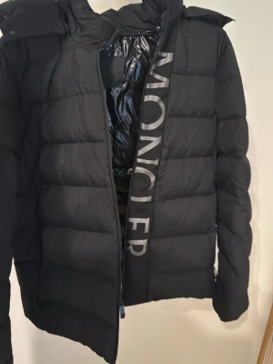  Moncler jacka - Säljer en svart pufferjacka från Moncler med stor logga på insidan av dragkedjan och Moncler-märke på ärmen. Jackan har huva, quiltad design och dragkedja framtill. Perfekt för kalla dagar och riktigt snygg till streetwear-stilen. Storlek S men passar både S och M