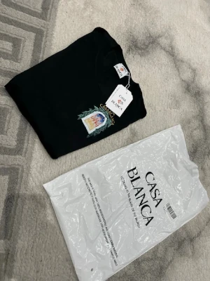 Casablanca T-shirt - Svart t-shirt från Casablanca Paris med färgglatt tryck på bröstet som visar en byggnad omgiven av gröna blad och texten 'CASABLANCA'. Rund hals och korta ärmar. T-shirten har en clean vibe med snygg grafik. Priset kan självklart diskuteras 😊 liten i storlek så rekommenderas till M (medium)