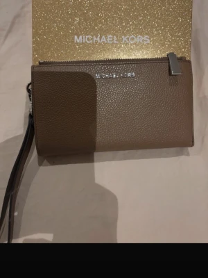 Brun plånbok från Michael Kors - Snygg brun plånbok från Michael Kors i strukturerat skinn. Plånboken har flera kortfack, dragkedja och handledsrem. Kommer i en glittrig guldfärgad presentask med logga. Perfekt för dig som vill ha både stil och funktion.