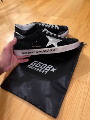 Golden Goose Super-Star sneakers svart/vit - Golden Goose Super-Star sneakers i svart mocka med vit stjärndetalj på sidan och handskriven text på sulan. Skorna har snörning, vit platt sula och svart/vita kontraster. Ikonisk streetstil och tillverkade i Italien. Kommer med originalpåse.