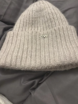 Beige stickad mössa från Carin Wester - Säljer en beige, ribbstickad mössa från Carin Wester med liten CW-logga framtill. Mössan är gjord i en mjuk blandning av polyester, akryl och angora, vilket gör den både varm och skön. Perfekt för kyliga dagar och enkel att matcha med olika outfits.