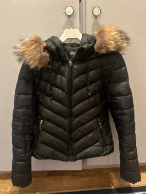 Svart pufferjacka  - Säljer en svart pufferjacka från Hollies med luva och fluffig päls. Jackan har dragkedja framtill, två fickor med dragkedja och snyggt quiltat mönster. Perfekt för kalla dagar och riktigt skön att bära.