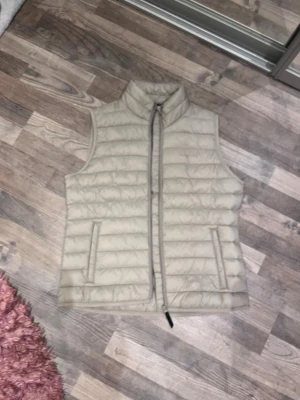 Beige dunväst från OCNOWKS M - Snygg beige dunväst från OCNOWKS i storlek M. Västen har quiltad design, hög krage och dragkedja framtill. Två fickor på sidorna och diskret logga på bröstet. Perfekt att slänga över en hoodie eller tröja för extra värme.